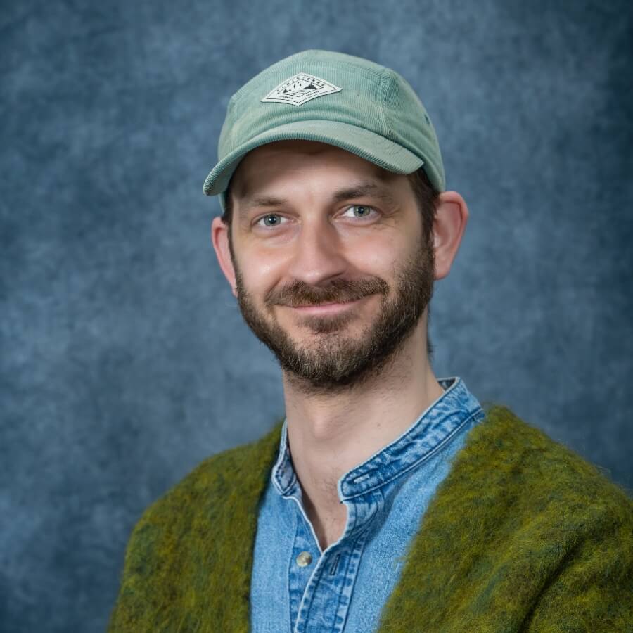 Mid-length shot of me - showing head and upper body. I am wearing a teal blue cord cap and a denim shirt over which is a fluffy green cardigan. In the background, a mottled blue backdrop.
