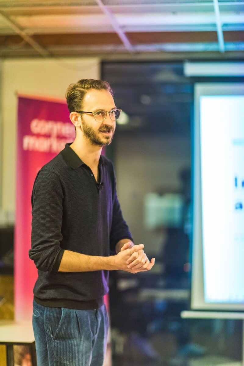 Shot in which I'm wearing a polo shirt, clear round rimmed glasses with a mid-length beard. My hands are clasped and mouth slightly open suggesting i'm mid way through a key point in my presentation. In the background there is a blurred pink and purple banner for the event.